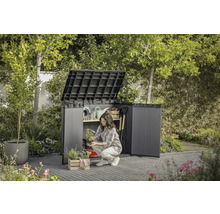 Gartenszene mit geöffnetem Geräteschrank, in dem eine Frau Gartenutensilien verstaut.