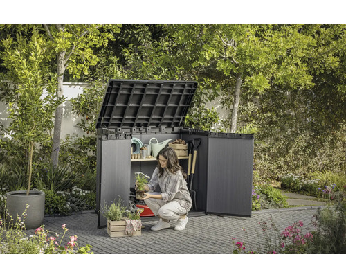 Gartenszene mit geöffnetem Geräteschrank, in dem eine Frau Gartenutensilien verstaut.