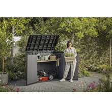 Gartenszene mit Gerätehaus, Gartengeräten und einer Frau, die eine Kiste mit Pflanzen hält.