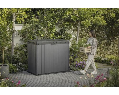 Gartenschrank aus Kunststoff im Garten mit Frau, die einen Korb mit Pflanzen trägt