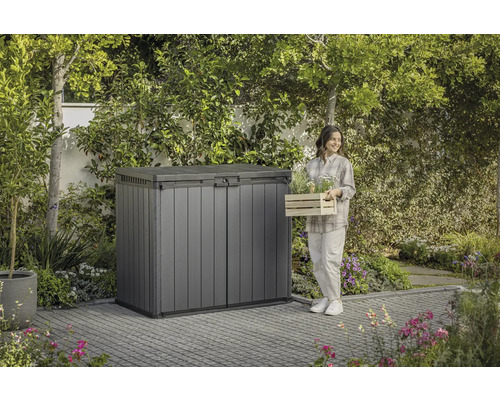 Gartenszene mit Gerätebox und einer Frau mit Pflanzenkiste.