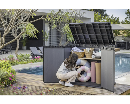 Szene im Garten mit einer Frau, die einen Geräteschrank aus Kunststoff mit zwei Regalböden öffnet, in dem sich Schwimmringe, Handtücher und Körbe befinden.