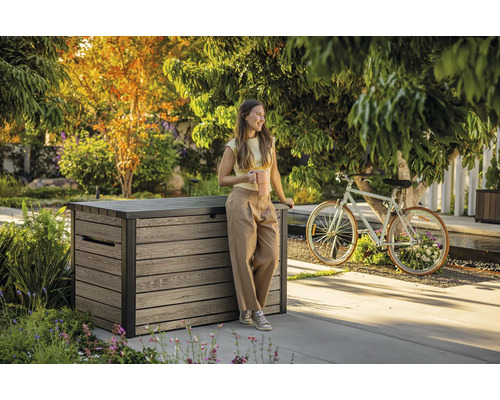 Szene im Garten mit einer Frau, die sich an eine Aufbewahrungsbox aus Holz lehnt und einen Kaffeebecher hält.