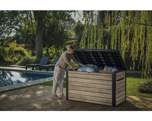 Eine Frau öffnet eine Gartentruhe auf einer Holzterrasse neben einem Pool und Garten.