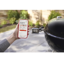 Une personne tient un smartphone avec l'application Weber Connect à côté d'un barbecue au charbon de bois.
