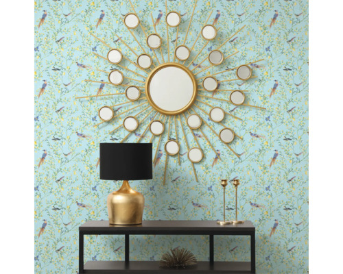Intérieur décoratif avec un miroir rond en forme de soleil, une lampe de table et une console devant un papier peint à motifs.