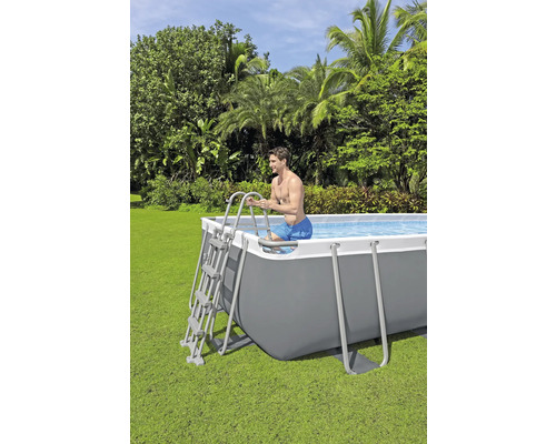 Un homme entre dans une piscine tubulaire rectangulaire dans un jardin en utilisant une échelle.