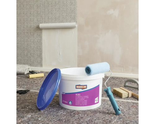 Szene mit einem Eimer Vliestapetenkleister, einer Farbwalze, einer Tapetenbürste und einer angefangenen Tapete an der Wand