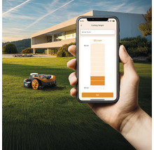 Scène avec un robot tondeuse et un smartphone affichant le réglage de la hauteur de coupe sur une pelouse devant une maison.