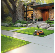 Un robot tondeuse tond la pelouse devant une maison moderne avec une façade en pierre et un grand jardin.