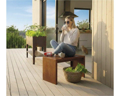 Szene auf einer Terrasse mit einem Hochbeet und einer Frau, die auf einem Hocker sitzt und ein Getränk genießt