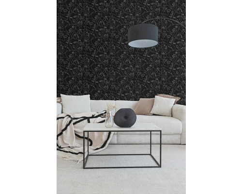 Wohnzimmer mit schwarzer Tapete mit weißen Gesichtern in Linienzeichnung, hellem Cordsofa und weißem Couchtisch mit schwarzem Metallgestell.