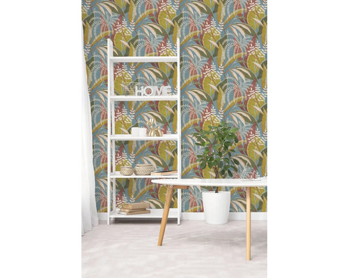 Vliestapete mit botanischem Blattmuster in Grün und Braun auf blauem Hintergrund, weißes Regal und Holztisch in hellem Raum.
