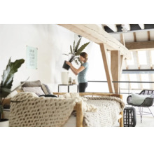 Helle, offene Wohnung mit Holzbalkendecke und einer Frau, die eine Zimmerpflanze in einen Übertopf umtopft.