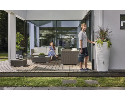 Ein moderner Terrassenbereich mit grauen Polyrattan-Gartenmöbeln, einer weißen Regentonne mit Pflanzgefäß und zwei Personen.