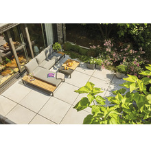 Terrasse moderne avec de grandes dalles grises, salon de jardin en L avec coussins gris et détails en bois, entouré de verdure.