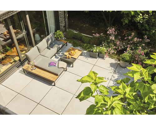 Terrasse moderne avec de grandes dalles grises, salon de jardin en L avec coussins gris et détails en bois, entouré de verdure.
