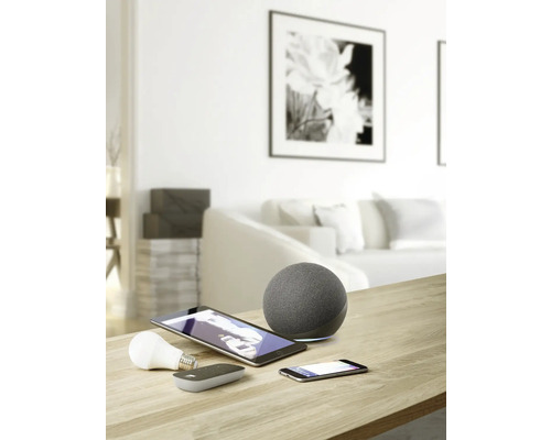 Aufnahme eines Wohnraums mit Smart Home Geräten wie Lautsprecher, Tablet, Smartphone und Leuchtmittel auf einem Tisch.