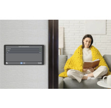 Panneau de commande Smart Home au mur affichant la consommation d'énergie, avec une femme sur un canapé dans un salon lumineux en arrière-plan.