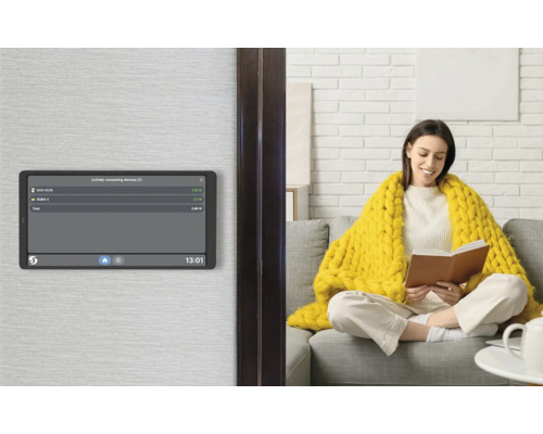 Panneau de commande Smart Home au mur affichant la consommation d'énergie, avec une femme sur un canapé dans un salon lumineux en arrière-plan.
