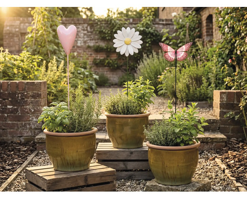 Drei Pflanztöpfe mit Kräutern und dekorativen Gartensteckern in Herz-, Blumen- und Schmetterlingsform in einem Garten.
