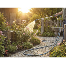 Eine Person bewässert Rosen mit einem Gartenschlauch auf einem gepflasterten Weg.