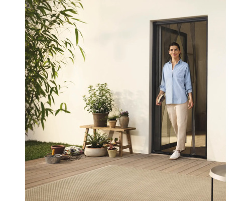 Frau betritt Terrasse durch Insektenschutztür, umgeben von Pflanzen und Gartenzubehör.