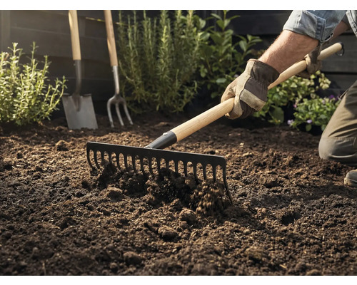 Gärtner harkt Erde mit einer Harke, im Hintergrund sind Spaten und Grabgabel zu sehen.