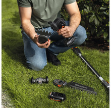 Une personne est accroupie sur la pelouse et tient une cisaille à gazon sans fil Husqvarna, avec différents accessoires et une batterie visibles sur l'image.
