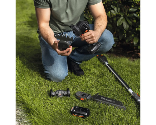 Une personne est accroupie sur la pelouse et tient une cisaille à gazon sans fil Husqvarna, avec différents accessoires et une batterie visibles sur l'image.