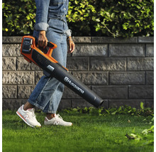 Une personne porte un souffleur de feuilles à batterie de marque Husqvarna dans un jardin.