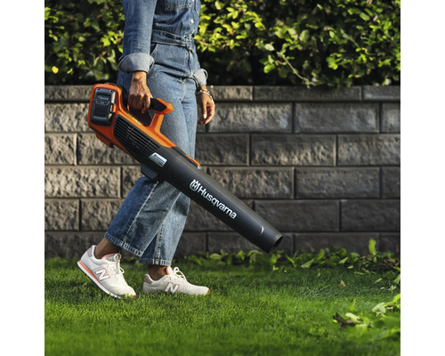 Une personne porte un souffleur de feuilles à batterie de marque Husqvarna dans un jardin.
