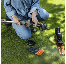 Une femme est accroupie sur la pelouse et assemble un taille-haie à batterie. Des accessoires sont posés sur la pelouse.