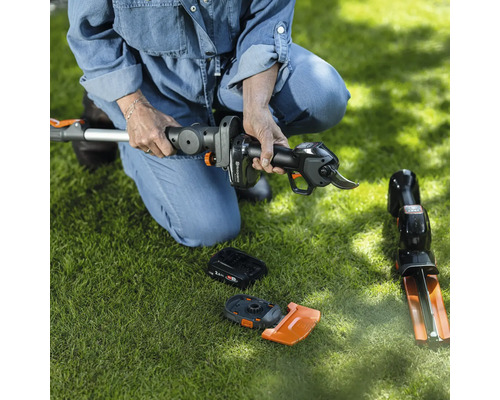 Une femme est accroupie sur la pelouse et assemble un taille-haie à batterie. Des accessoires sont posés sur la pelouse.