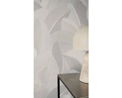 Vliestapete mit großflächigem, strukturiertem Blattmuster in neutralen Grautönen an einer Wand hinter einer modernen Tischlampe.