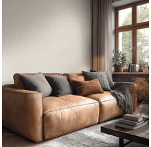 Helles Wohnzimmer mit großem braunem Ledersofa, verschiedenen Kissen, grauer Decke, strukturierter Tapete und Fenster aus Holz.