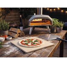 Photo en extérieur d'un four à pizza Ooni avec pizza, pelle à pizza et ingrédients sur une table en bois.