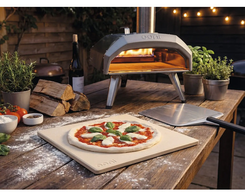 Photo en extérieur d'un four à pizza Ooni avec pizza, pelle à pizza et ingrédients sur une table en bois.