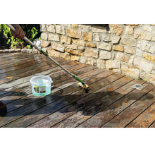 Nettoyage d'une terrasse en bois avec une brosse à long manche et du gel dégriseur Osmo devant un mur en pierre.