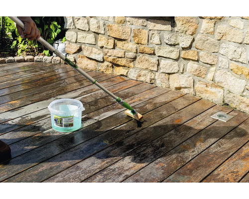 Nettoyage d'une terrasse en bois avec une brosse à long manche et du gel dégriseur Osmo devant un mur en pierre.