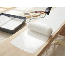 Une planche en bois est peinte avec un rouleau à peinture et de la peinture blanche, un bac à peinture, des pinceaux et du ruban de masquage sont prêts.