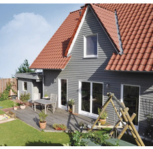 Maison avec revêtement de façade horizontal gris, toit en tuiles rouges et terrasse en bois avec table, chaises et plantes en pot à côté d'une pelouse.