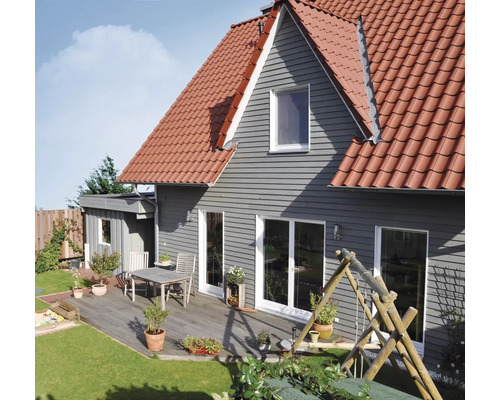 Maison avec revêtement de façade horizontal gris, toit en tuiles rouges et terrasse en bois avec table, chaises et plantes en pot à côté d'une pelouse.