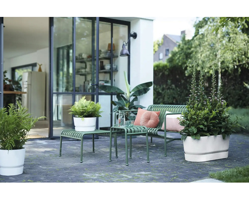 Moderne Terrasse mit grüner Gartenbank aus Metall, Beistelltischen und weißen Blumentöpfen vor einer Glasfront.