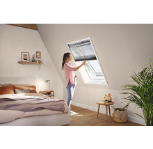 Eine Frau bedient ein dunkles Plissee an einem weißen Dachfenster in einem hellen, modernen Schlafzimmer mit Dachschräge.