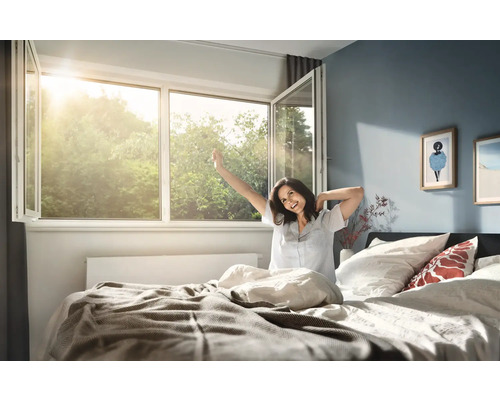Grille de protection anti-pollen Tesa® 1,5x1,8 m Chambre lumineuse avec fenêtre en plastique grande ouverte laissant entrer le soleil et une personne s'étirant dans son lit au réveil.