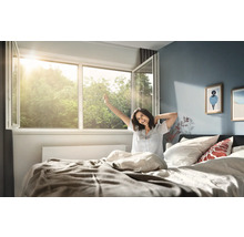 Helles Schlafzimmer mit weit geöffnetem Kunststofffenster, durch das Sonnenlicht fällt, und eine Person im Bett, die sich beim Aufwachen streckt.