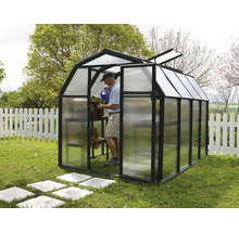 Kleines Gewächshaus in Scheunenform mit schwarzem Rahmen und Polycarbonat-Paneelen im Garten. Ein Mann gießt im Inneren Pflanzen.