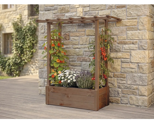 Jardinière en bois avec treillis intégré et pergola, plantée de fleurs et de tomates devant un mur en pierre naturelle.