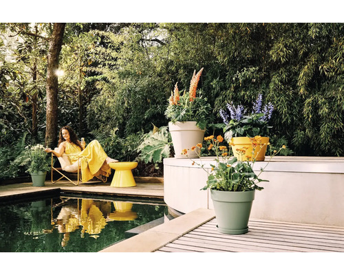 Femme en robe jaune sur une terrasse en bois au bord de la piscine, entourée de pots de fleurs et de plantes luxuriantes dans un jardin lumineux.